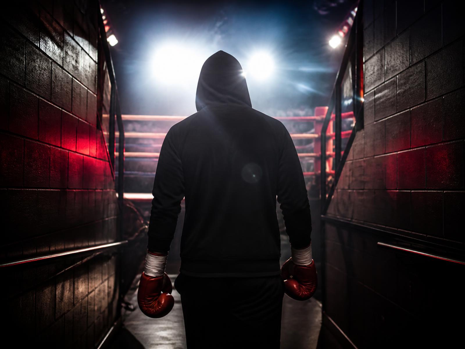 Hooded fighter walking a dark tunnel toward a lit boxing ring